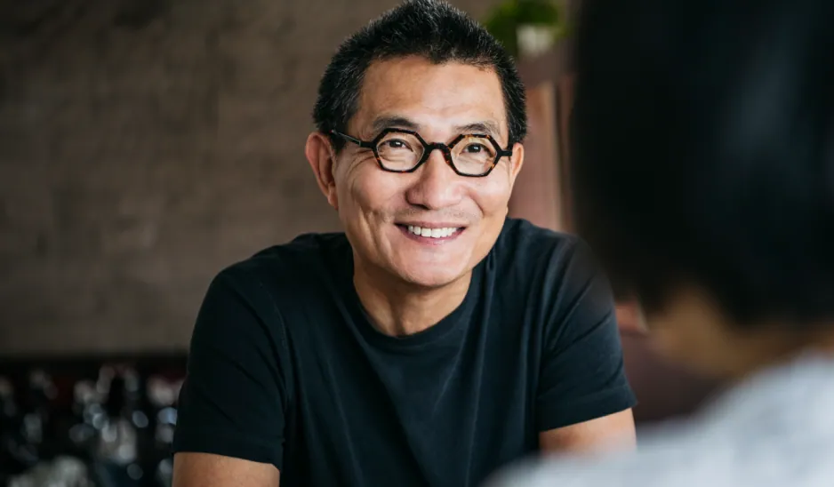 The man wearing glasses smiling as he converses, with a solid brick wall in the background