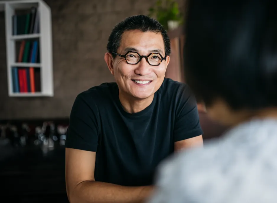 The man wearing glasses smiling as he converses, with a crooked, surreal-looking bookshelf in the background